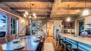 Un espace repas avec une table en bois dressée pour quatre personnes, un escalier menant à une mezzanine et une cuisine dotée d’un grand îlot central.