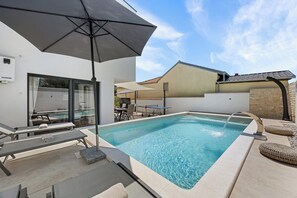 Piscina com espreguiçadeiras e um guarda-sol, rodeada por um edifício com telhado em azulejos.
