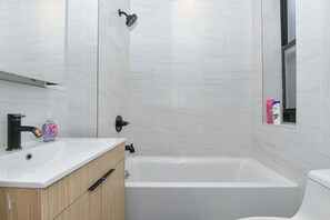 A modern bathroom with a white bathtub, a wooden vanity, a black faucet, and a showerhead. - Chelsea Charm : 3BR Retreat Near High Line (New York)