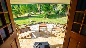 Un patio exterior con suelo de piedra, dos sillas de madera, una mesita y un par de chanclas sobre una esterilla.