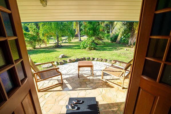 An outdoor patio with stone flooring, two wooden chairs, a small table, and a pair of flip-flops on a mat. - Fare Opuhi (Moorea-Maiao)