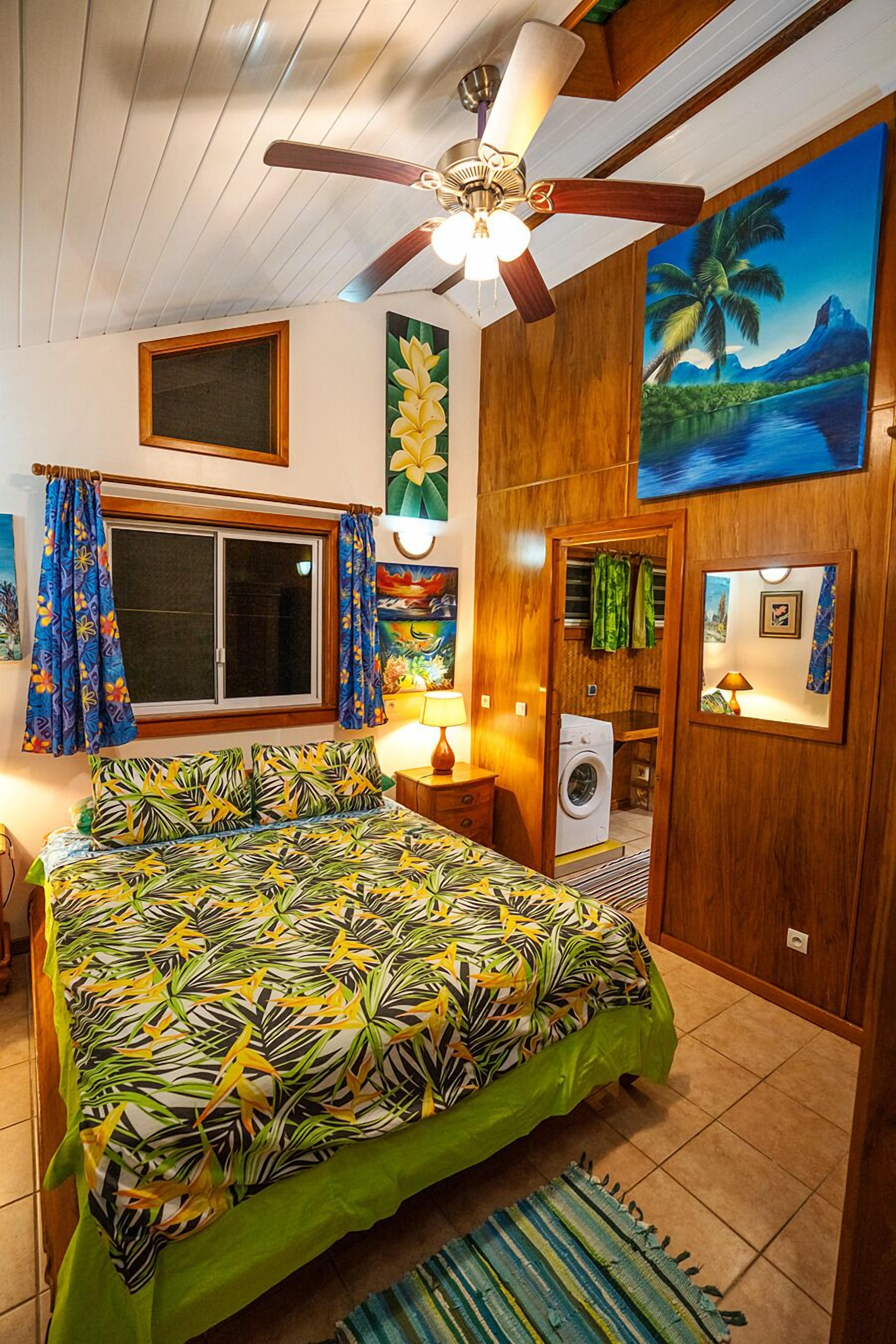 A bedroom with a bed, a ceiling fan, a window with curtains, a washing machine, and a painting on the wall.