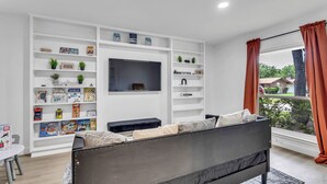 Una sala de estar moderna con un sofá negro, estanterías blancas llenas de libros infantiles, un televisor y una ventana con cortinas.