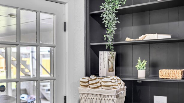 Una habitación con una puerta blanca, una cesta de toallas a rayas, una planta en maceta y un estante con libros y un letrero.