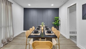 Comedor moderno con una mesa larga dispuesta para ocho comensales, rodeada de sillas de mimbre, frente a una pared oscura con una lámpara en el techo.