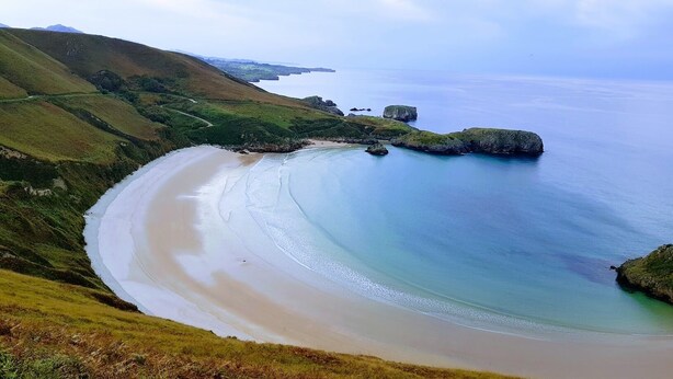 A coastal landscape with a curved sandy beach, grassy hills, and rocky outcrops.