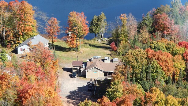 Une cabane avec une allée en gravier, entourée d’arbres aux couleurs automnales et bordée d’une étendue d’eau au loin.