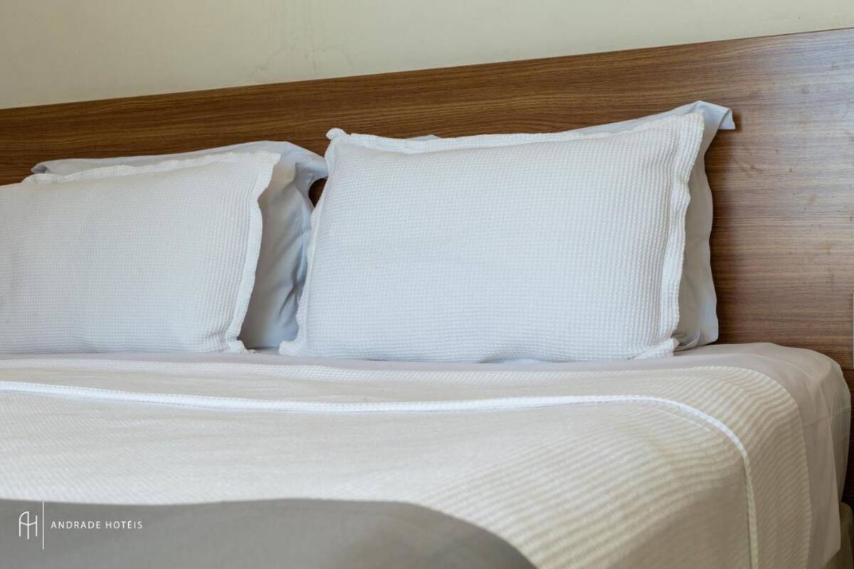 Un letto ben rifatto con biancheria e cuscini bianchi, una testiera in legno e il logo di Andrade Hotels.