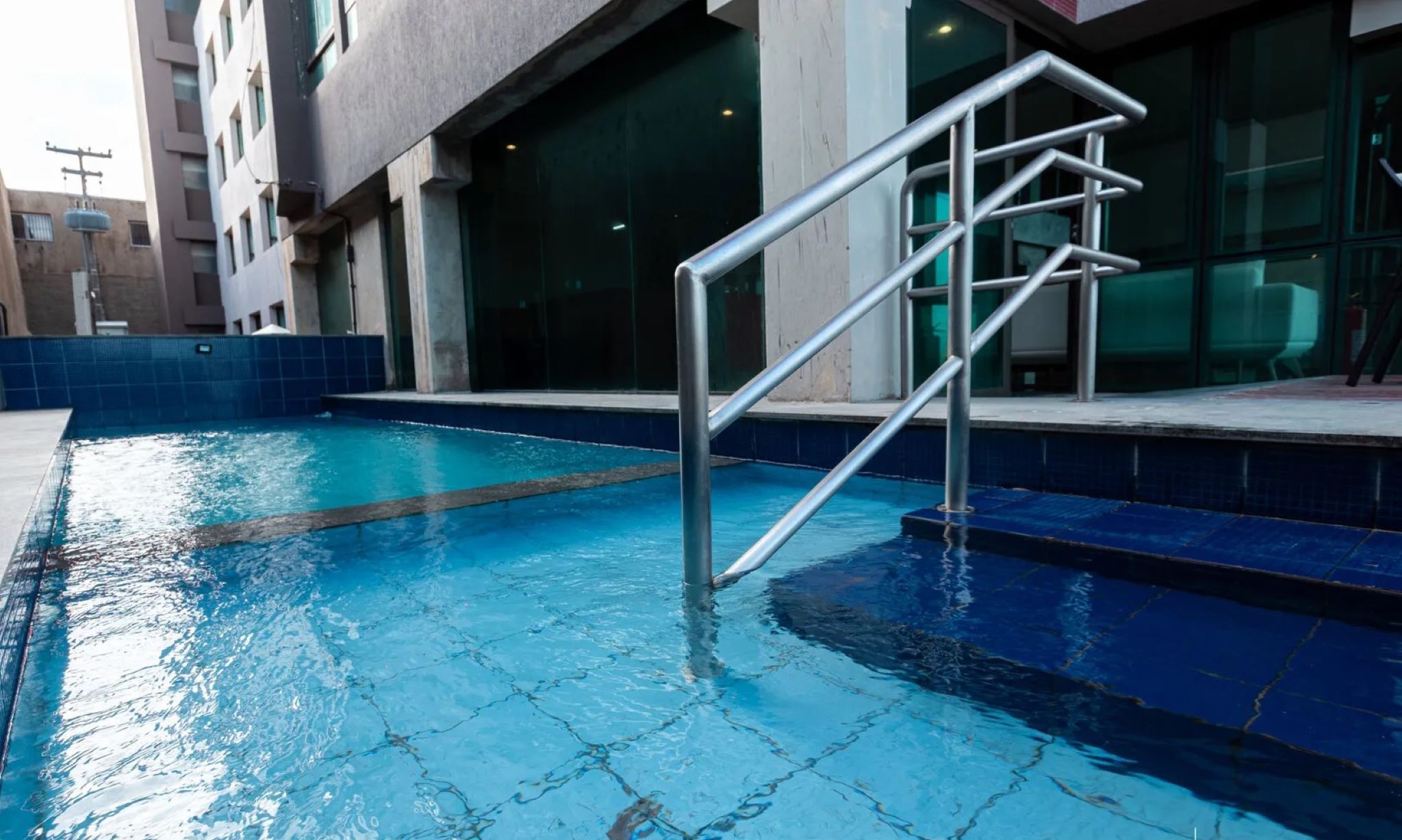 Una piscina con una escalera metálica que da al agua, rodeada por un solárium con baldosas y un edificio con grandes ventanales.