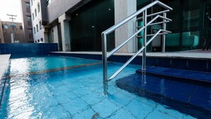 En swimmingpool med en metalstige ned i vandet, omgivet af en fliseterrasse og en bygning med store vinduer.