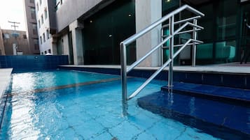 Una piscina con una escalera metálica que da al agua, rodeada por un solárium con baldosas y un edificio con grandes ventanales.
