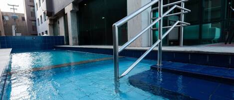 Una piscina con una escalera metálica que da al agua, rodeada por un solárium con baldosas y un edificio con grandes ventanales.