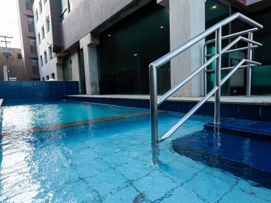 Una piscina con una escalera metálica que da al agua, rodeada por un solárium con baldosas y un edificio con grandes ventanales.