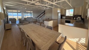 Un espai d'estar ampli amb una gran taula de dinar de fusta, cadires de vimetla i vista a la platja a través de grans finestres.