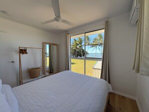 A bedroom with a bed, a ceiling fan, a window with a beach view, and a wooden floor.