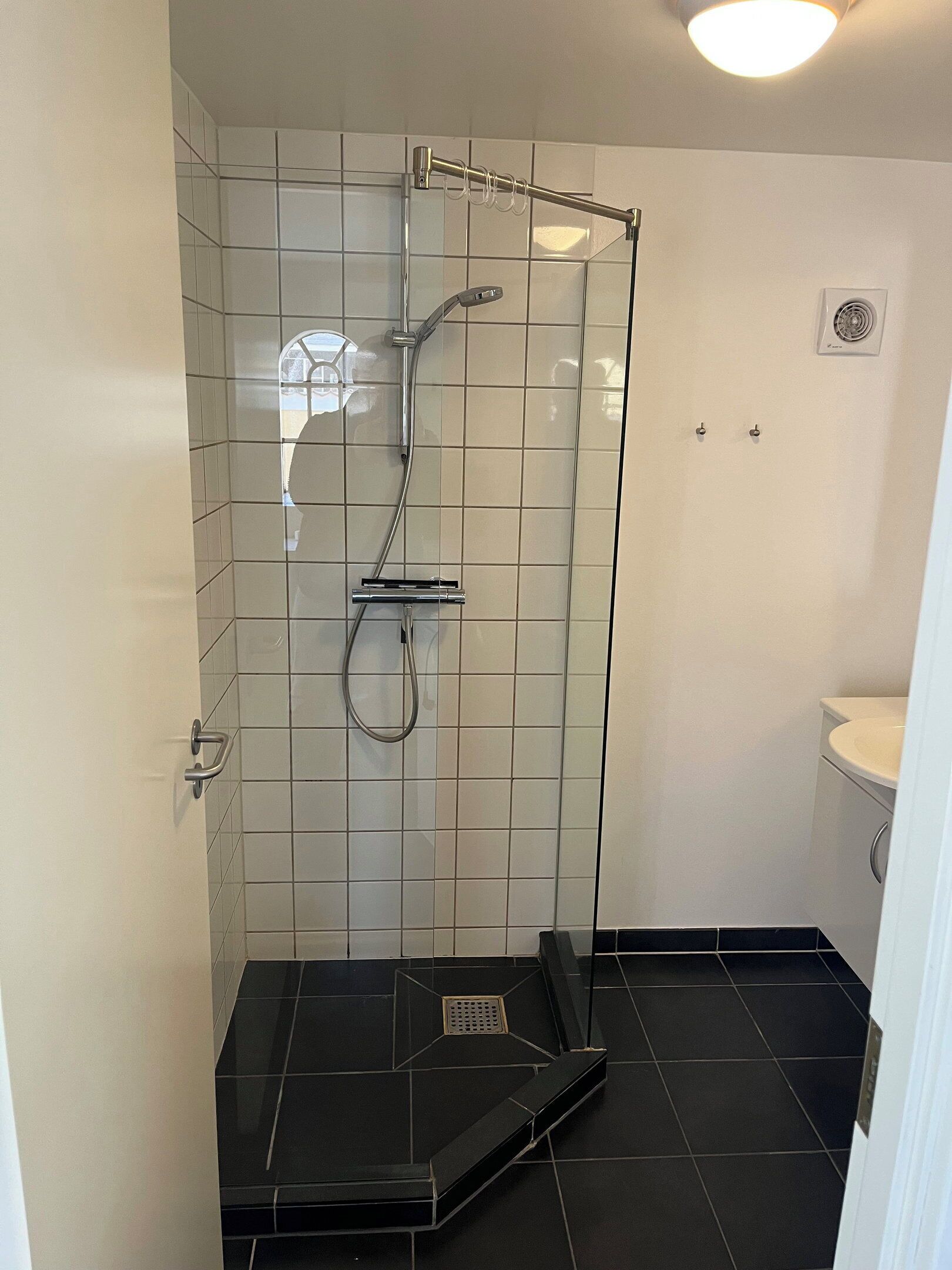 A shower with a glass door, a white tiled wall, and a black tiled floor.