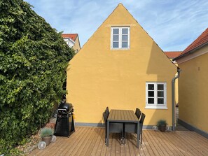 A yellow building with a triangular roof, a window, and a table with chairs in the foreground.