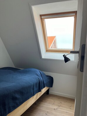 Une petite chambre avec un lit simple, un plafond en pente, un puits de lumière et une lampe de bureau.