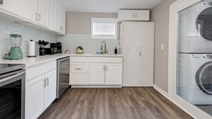 A compact kitchen with white cabinets, a built-in dishwasher, a countertop with a blender, and a large window with blinds.