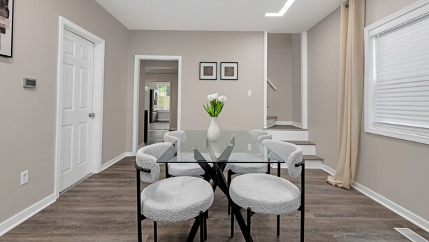 A modern dining area with a glass table, six upholstered chairs, a vase with flowers, and a staircase in the background.