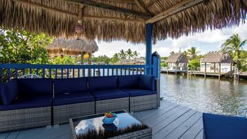Relax in a serene outdoor seating area with a thatched roof, adjacent to a calm body of water and surrounded by a blue railing.