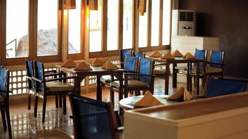 Un restaurante con mesas de madera y sillas azules, amplias ventanas con vistas a rocas y un equipo de aire acondicionado en la pared.