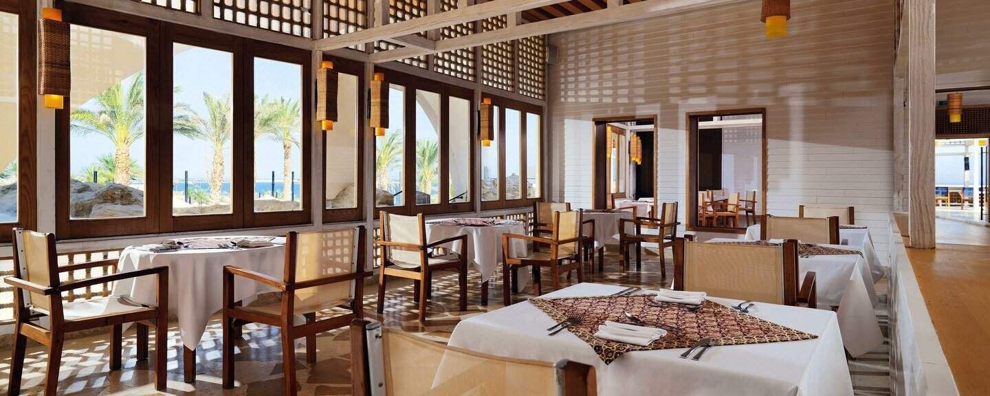 Un restaurant doté de nombreuses tables et chaises, de grandes fenêtres offrant une vue sur les palmiers et la mer, et d’une structure de plafond en bois.