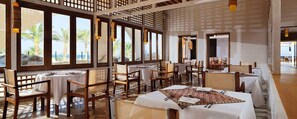 A restaurant with multiple tables and chairs, large windows with a view of palm trees and the sea, and a wooden ceiling structure.