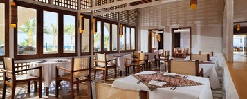 Un restaurante con varias mesas y sillas, amplias ventanas con vistas a palmeras y al mar, y una estructura de techo de madera.