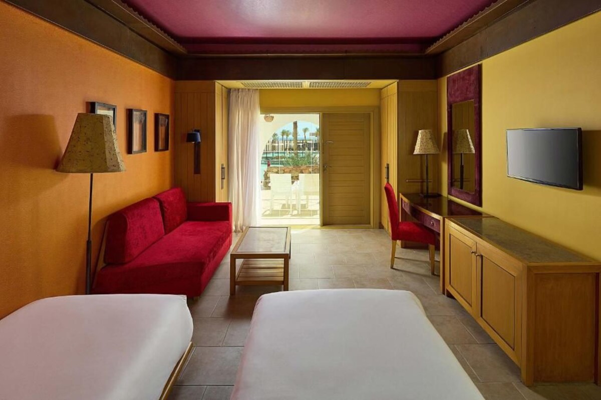 A hotel room with two beds, a red sofa, a wooden desk, a TV, and a view of a pool area.