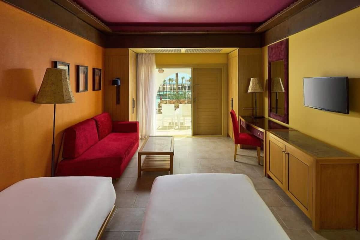 A hotel room with two beds, a red sofa, a wooden desk, a TV, and a view of a pool area.
