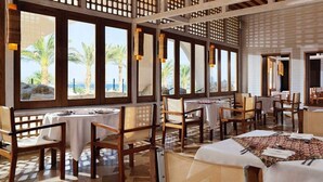 Ein Restaurant mit mehreren Tischen und Stühlen, großen Fenstern mit Blick auf Palmen und Meer, und einer Holzbalkendecke.