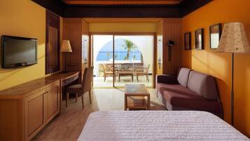 A hotel room with a bed, a desk with a chair, a sofa, a TV, and a view of the sea through a glass door.