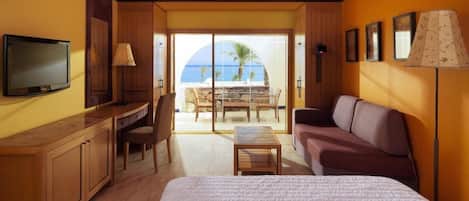 A hotel room with a bed, a desk with a chair, a sofa, a TV, and a view of the sea through a glass door.