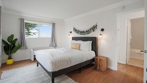 Ein Schlafzimmer mit Bett, Nachttisch, Lampe, Topfpflanze und Blick ins Badezimmer.