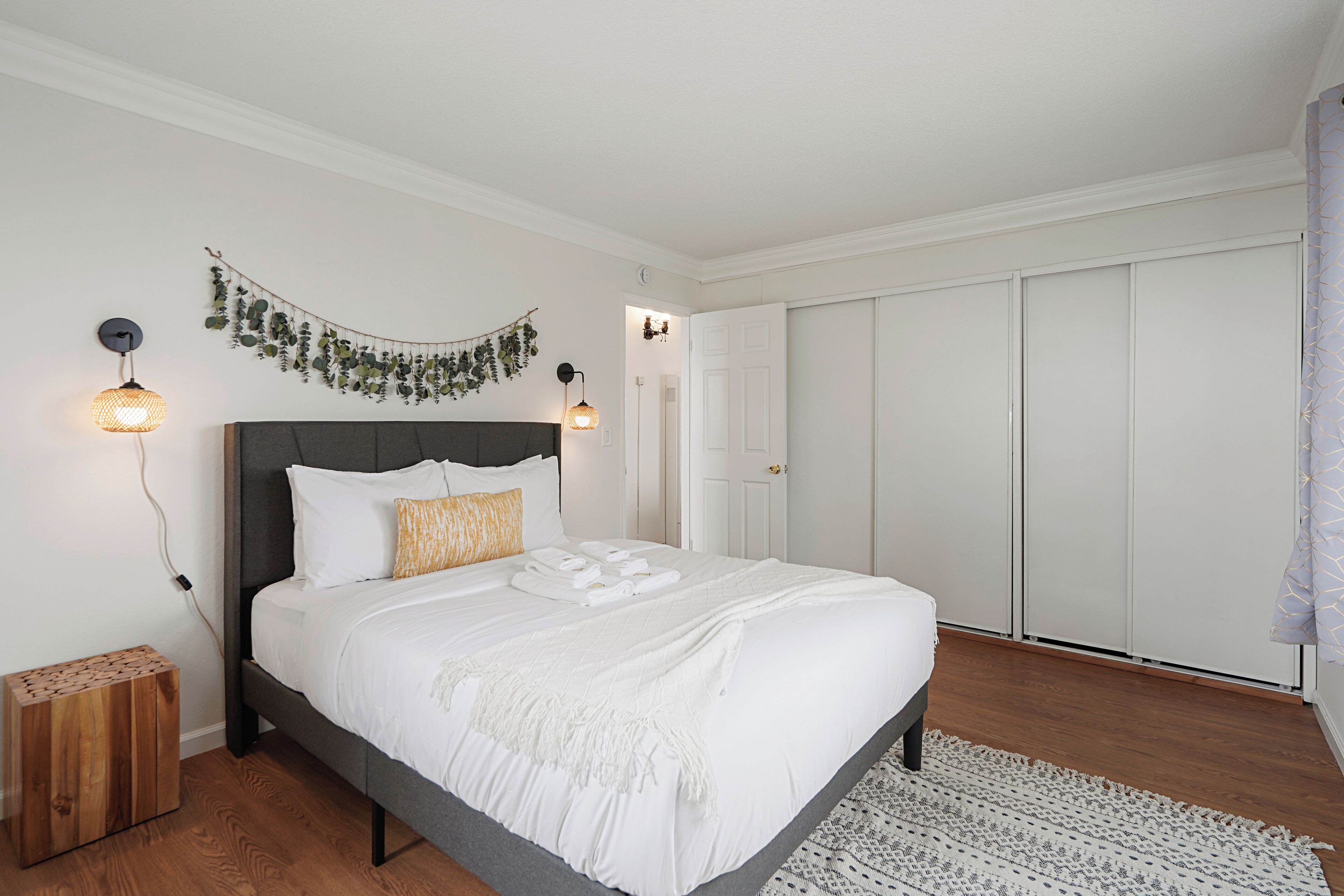 A neatly made bed with white linens, a grey headboard, and a decorative wall hanging. There is a wooden nightstand with a patterned lamp beside the bed.