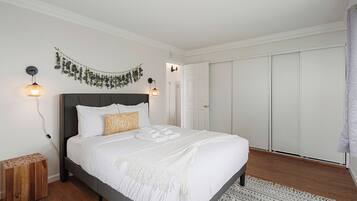 A neatly made bed with white linens, a grey headboard, and a decorative wall hanging. There is a wooden nightstand with a patterned lamp beside the bed.