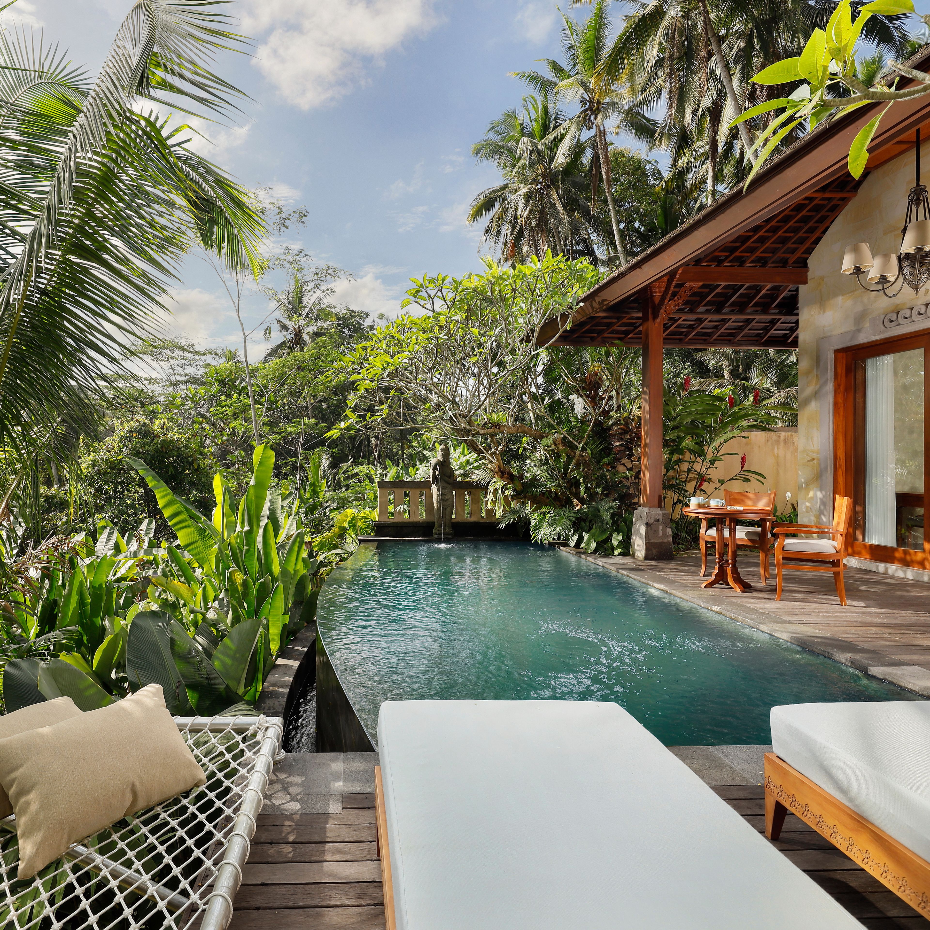 Zona junto a la piscina con una silla de descanso, una mesa de madera con sillas, y vistas a la piscina rodeada de vegetación tropical.