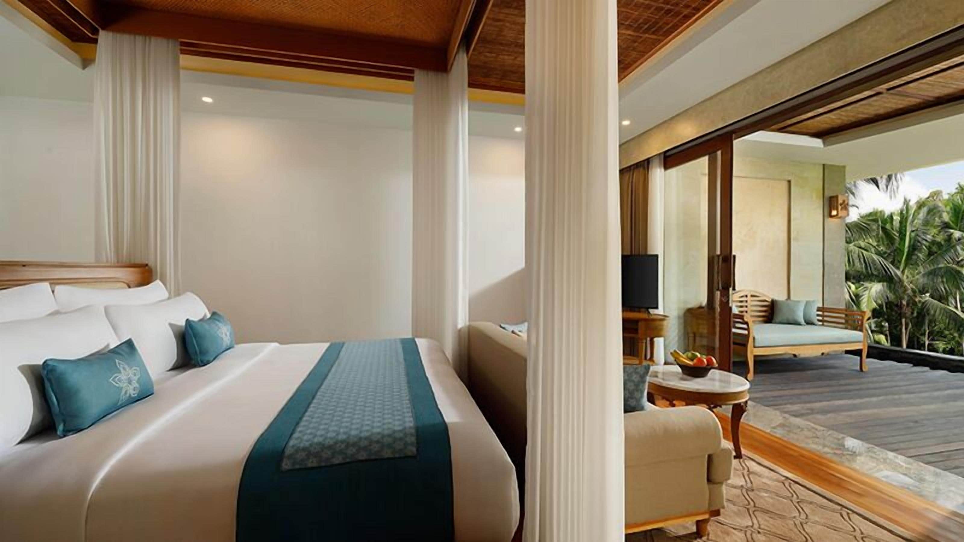 Une chambre d’hôtel avec un grand lit, une petite table sur laquelle se trouve un saladier de fruits, et une vue sur un balcon agrémenté d’un canapé et de palmiers.