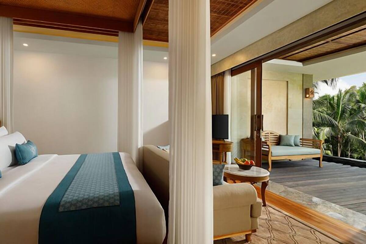 A hotel room with a large bed, a small table with a bowl of fruit, and a view of a balcony with a sofa and palm trees.