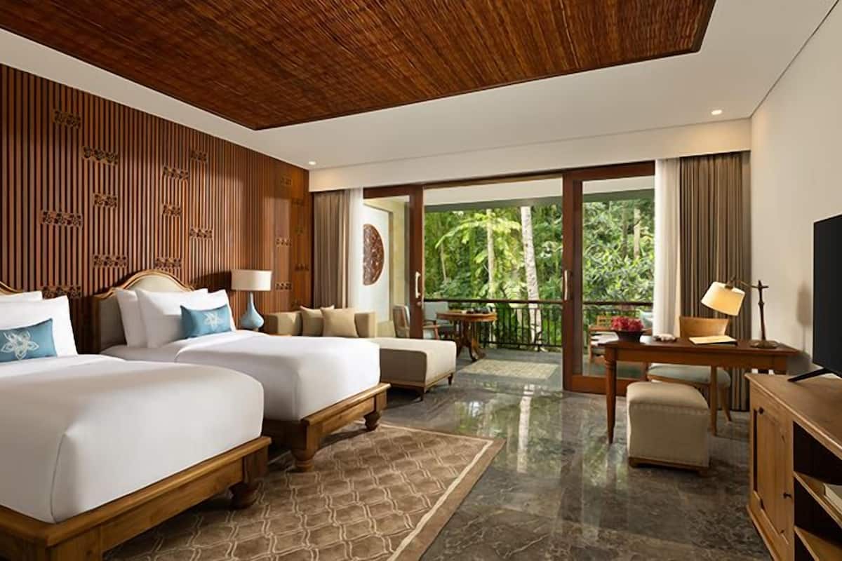 A hotel room with two beds, a balcony with a view of greenery, a desk with a TV, and a wooden ceiling.
