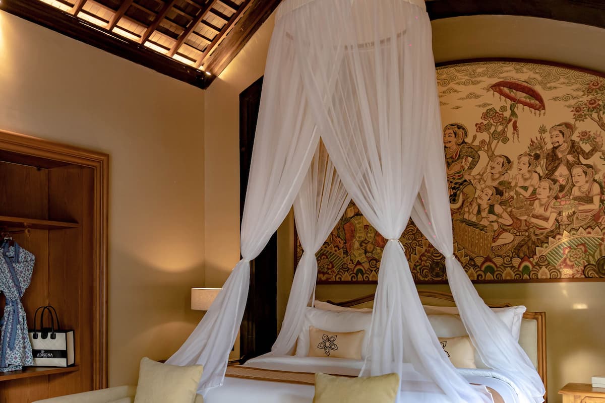 A room with a bed under a canopy, a sofa, a small table with a bowl of fruit, and a wall with a tapestry.