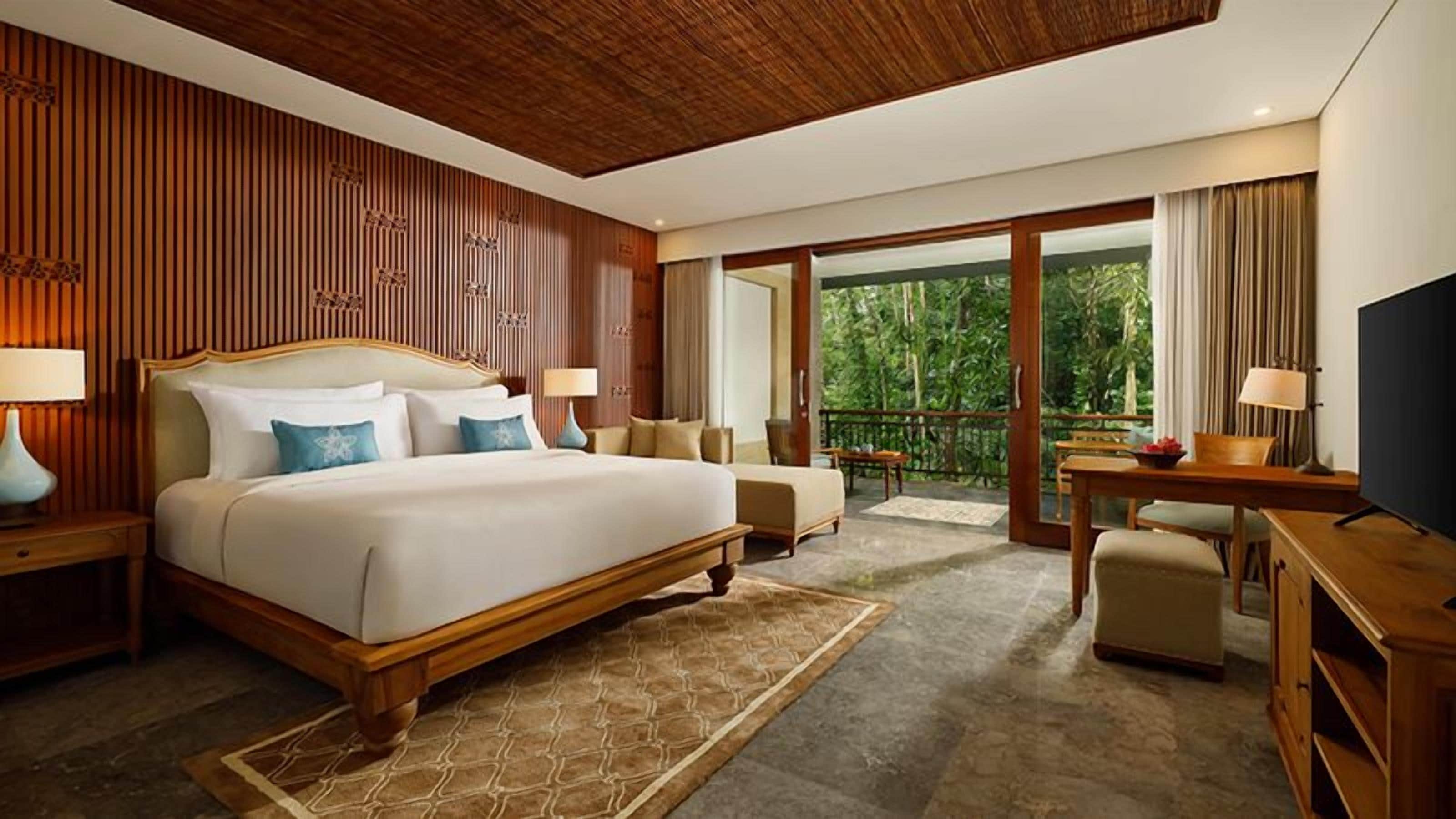 Habitación de hotel con una cama grande, un escritorio, televisión y balcón con vistas a zonas verdes.
