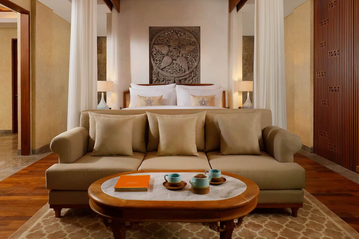 A hotel room with a sofa, a coffee table with a book and cups, a bed with pillows, and a decorative wall piece.