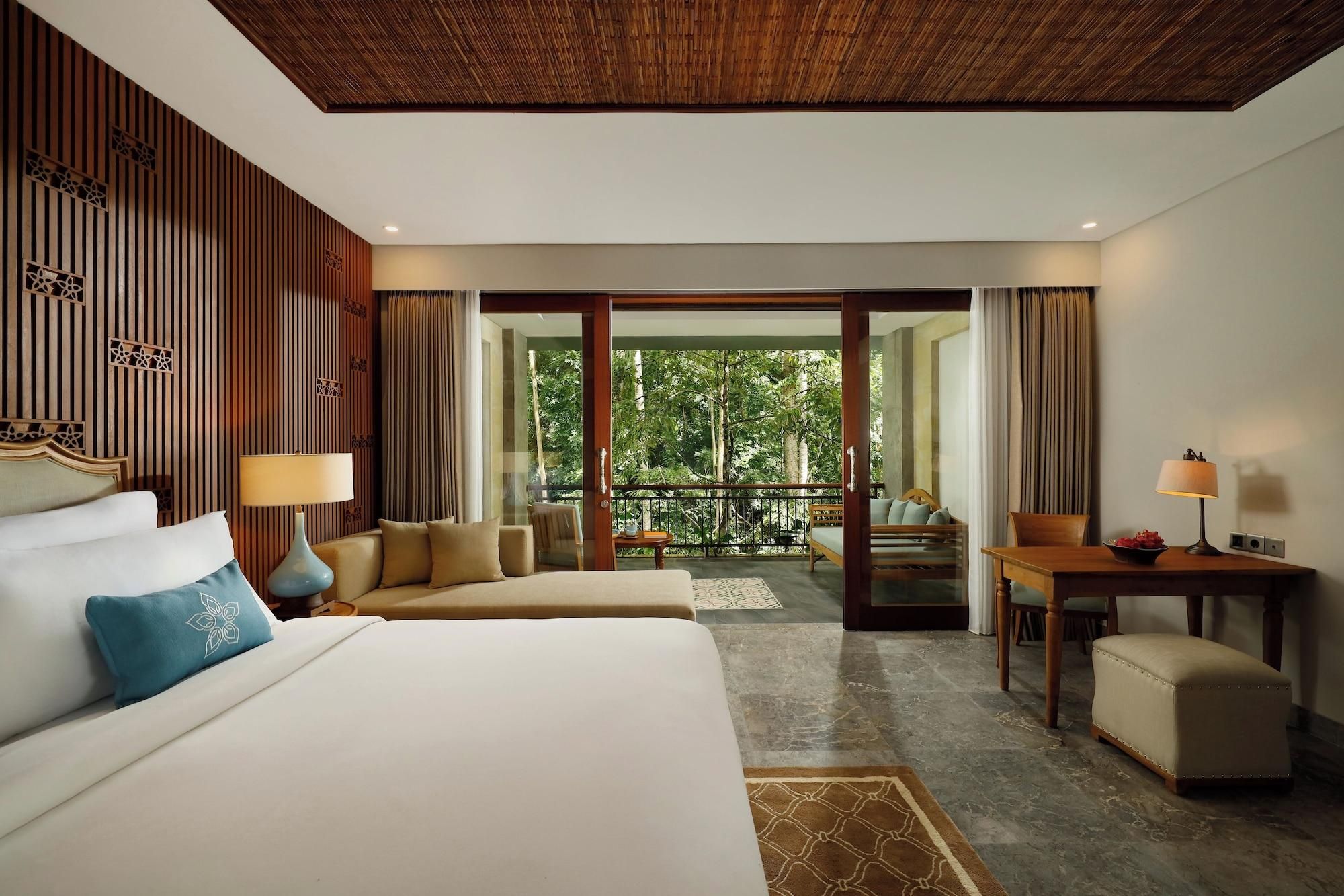 A hotel room with a large bed, a sofa, a table, and a view of a balcony with trees.