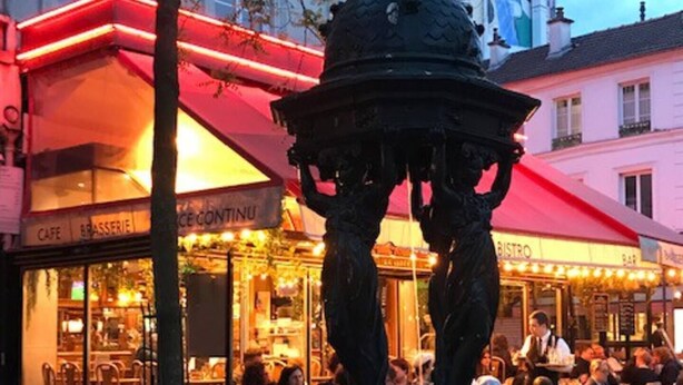 Un espace café en plein air avec des tables et des chaises, une fontaine décorative et, au fond, un bâtiment surmonté d’un auvent rouge.