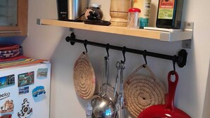 Une cuisine avec une étagère où sont posés un faitout, une bouteille et un bocal. En dessous, on peut voir des casseroles et des poêles suspendues.