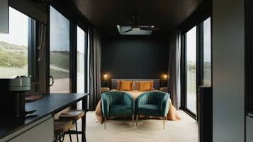 A bedroom with a bed, a desk, and a chair, large windows, and a view of the outdoors.