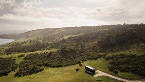 An aerial view of a hilly landscape with a road leading to a small building, likely a cabin or lodge, surrounded by dense vegetation and overlooking a body of water. - Cabn Kangaroo Island (Ironstone)