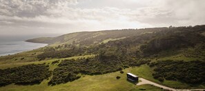 Vista aerea di un paesaggio collinare con una strada che conduce a un piccolo edificio, probabilmente una baita o un lodge, immerso nella vegetazione e con vista su un corpo d'acqua.